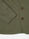Close-up of a light olive twill Bakers Jacket on a white surface. The jacket features brown buttons and visible stitching details.