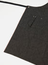 Close-up of a Black Selvedge Denim Apron on a white background. The apron has a black strap and red selvedge detail. Features pockets with button closures.